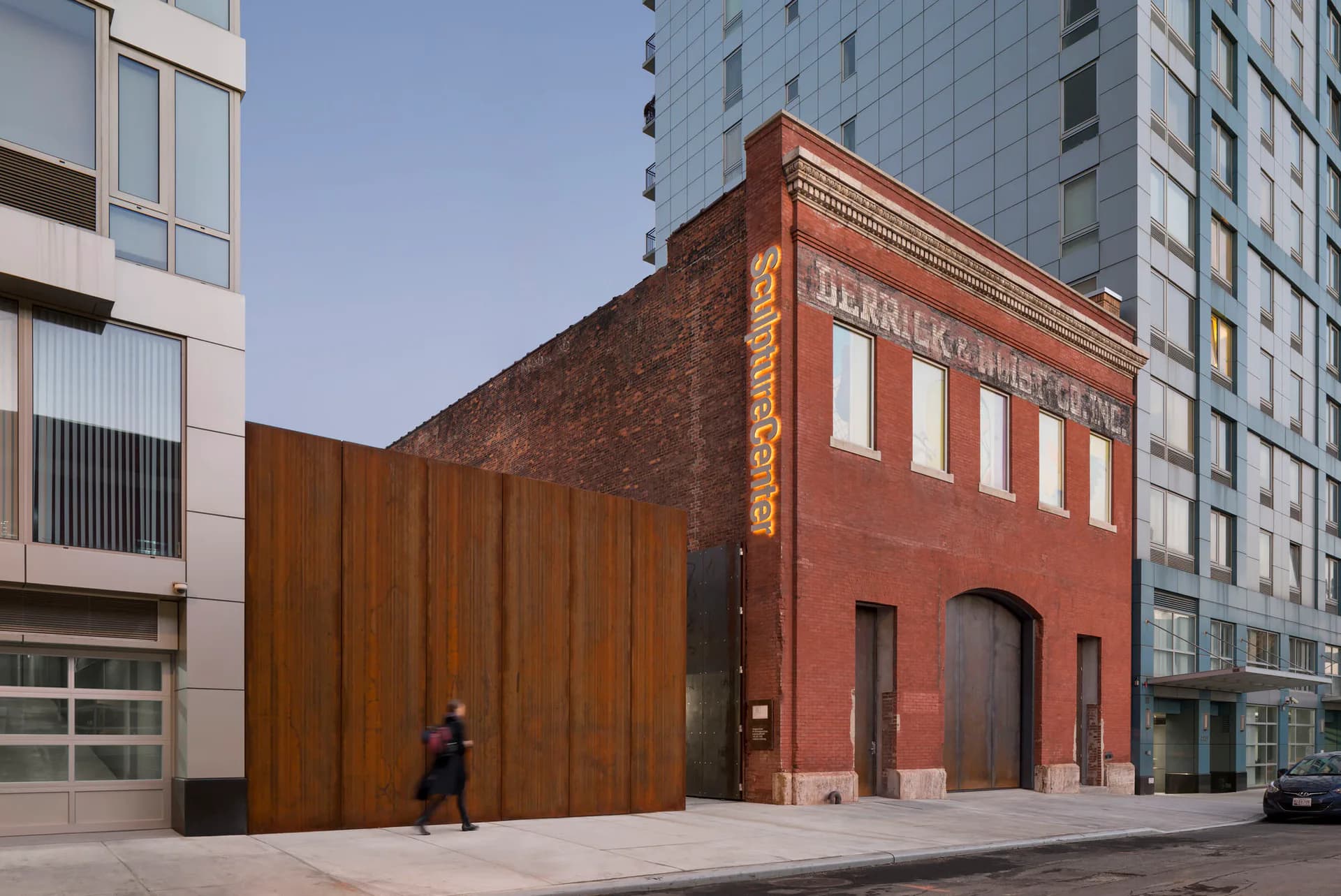 Exterior courtyard of Sculpture Center with red brick building facade, large wooden panel wall, and modern glass buildings adjacent.