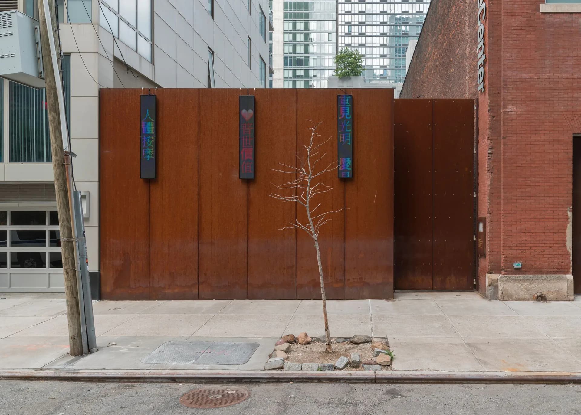 Courtyard view of SculptureCenter featuring a large rust-colored steel wall with vertical blue panel details and a young sapling tree in the center, surrounded by modern buildings.