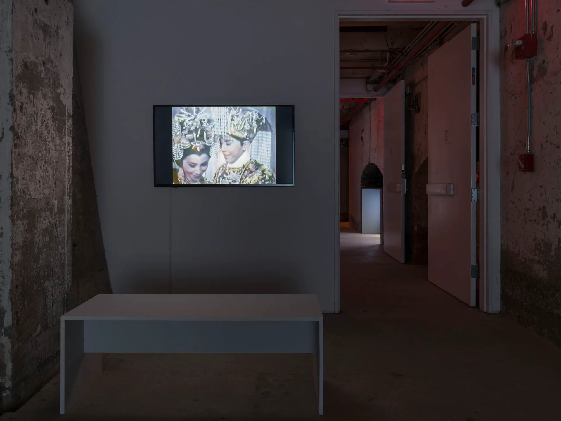 Interior gallery corridor at SculptureCenter with exposed brick walls, gray-painted sections, video monitor mounted on wall displaying artwork, and multiple doorways leading deeper into space.