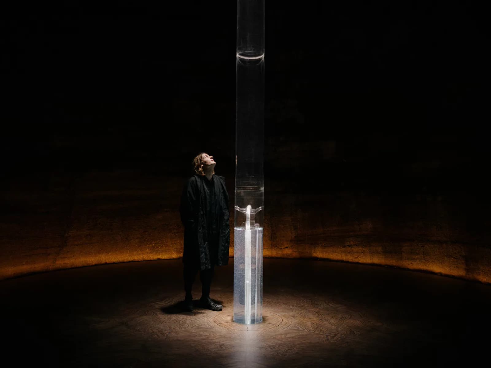 Installation artwork featuring a figure in dark clothing standing beside an illuminated vertical rectangular form against a dark background with warm orange lighting.