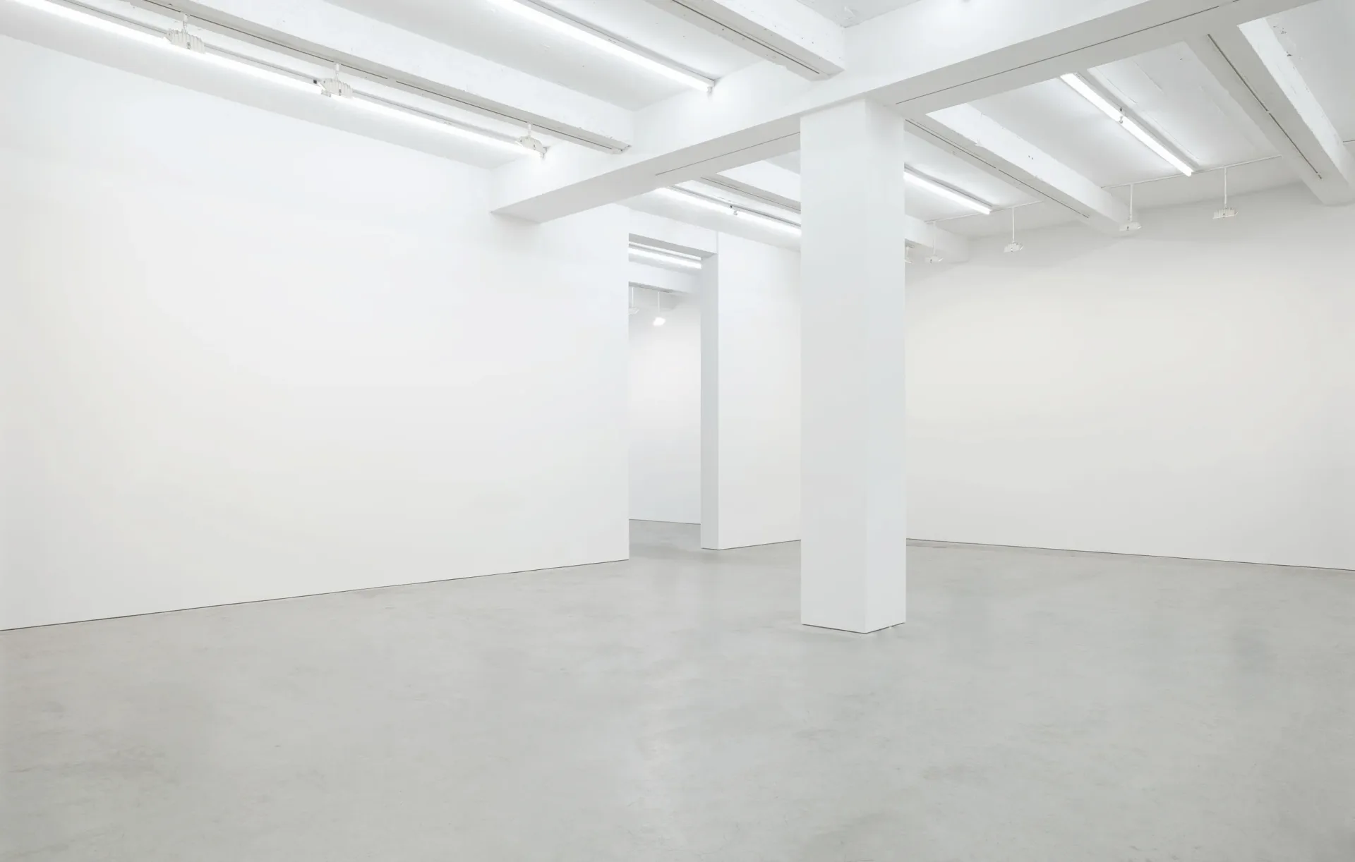 Bright, minimalist gallery interior with white walls, polished concrete floor, fluorescent ceiling lights, and a central support column.