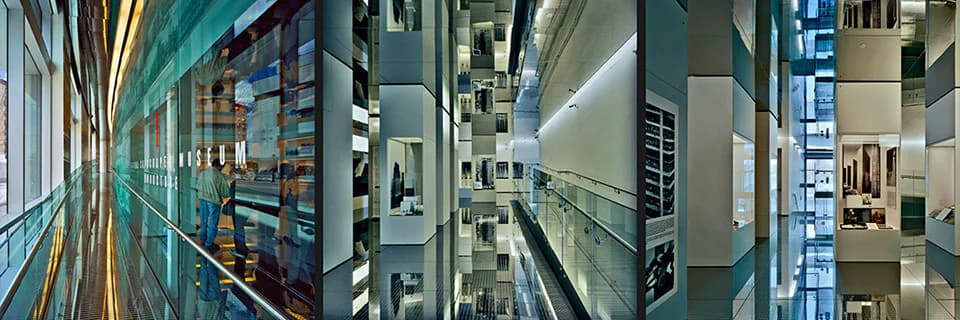 Skyscraper Museum gallery corridor with teal-colored upper walls, display cases, glass barriers, and overhead lighting creating a modern exhibition space.