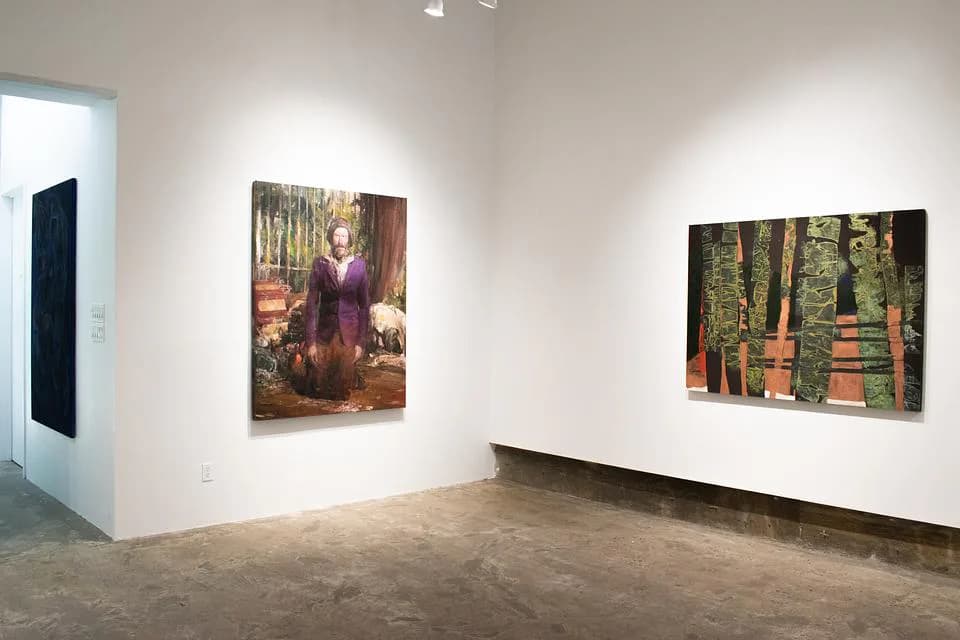Gallery interior with white walls, concrete floor, and several paintings on display including a portrait and a forest-themed artwork