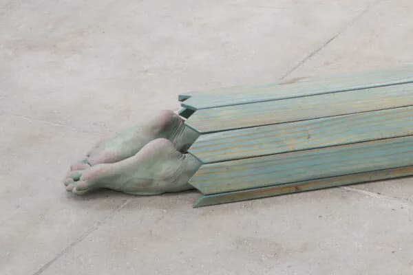 Sculpture of human legs and feet in pale green-blue painted wood or metal, positioned horizontally on concrete floor.