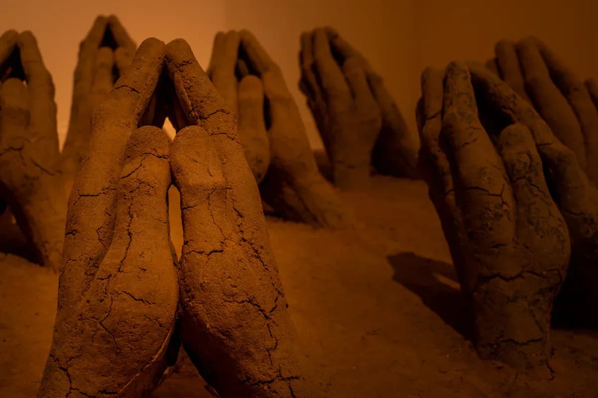 Sculptural installation of clay or ceramic hand forms arranged on a sandy surface under golden ambient lighting, creating dramatic shadows.