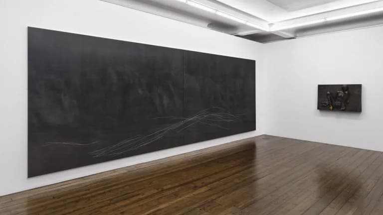 Large-scale abstract charcoal or graphite drawing on black wall panel with gestural marks, displayed in white gallery space with wooden floor.