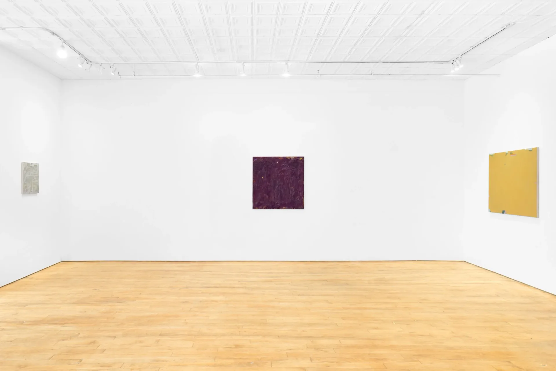 Empty gallery space with wooden floor and three small framed artworks—burgundy, pale gray, and mustard yellow—on white walls.