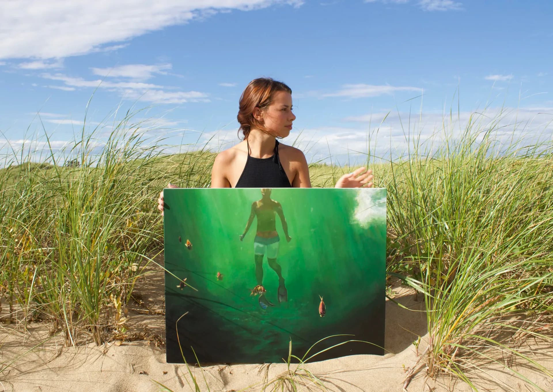 Framed painting held by woman in beach dunes showing underwater green portrait against cloudy blue sky and water.
