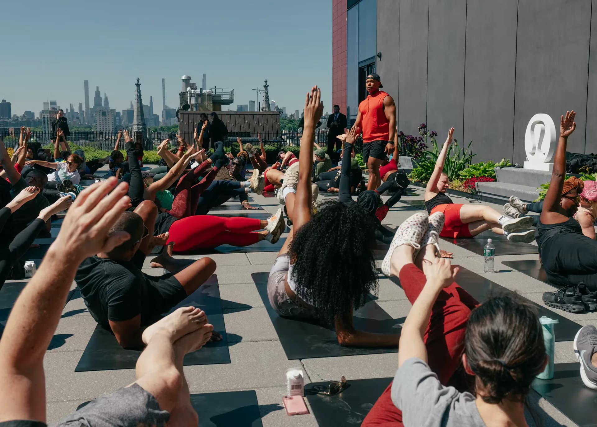 Outdoor rooftop gathering with crowd of people in yoga poses on mats with Manhattan skyline and buildings in background.