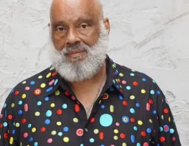 Portrait photograph of a man with white beard wearing a colorful polka-dot shirt against a marble background.