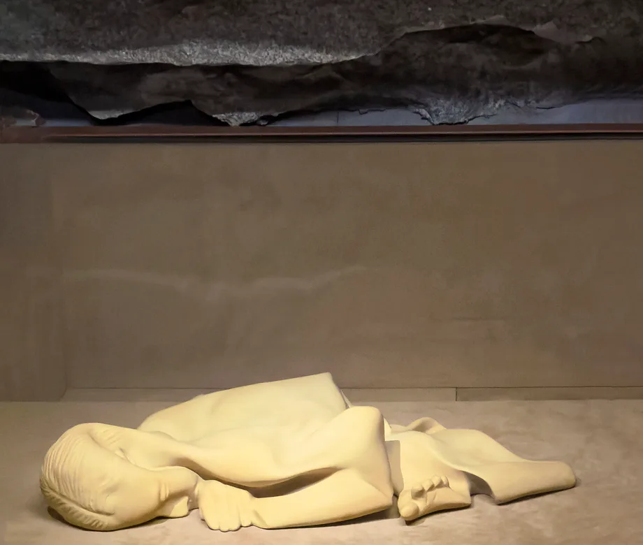 Sculptural installation of cream-colored crumpled fabric forms on beige floor beneath dark ceiling element, creating organic, abstract shapes.