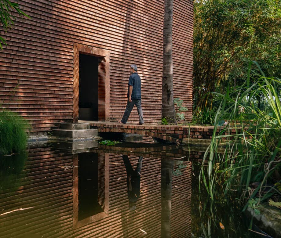 Exterior of brick gallery building with horizontal wooden siding, dark doorway, figure walking on bridge over reflecting pool, surrounded by vegetation.