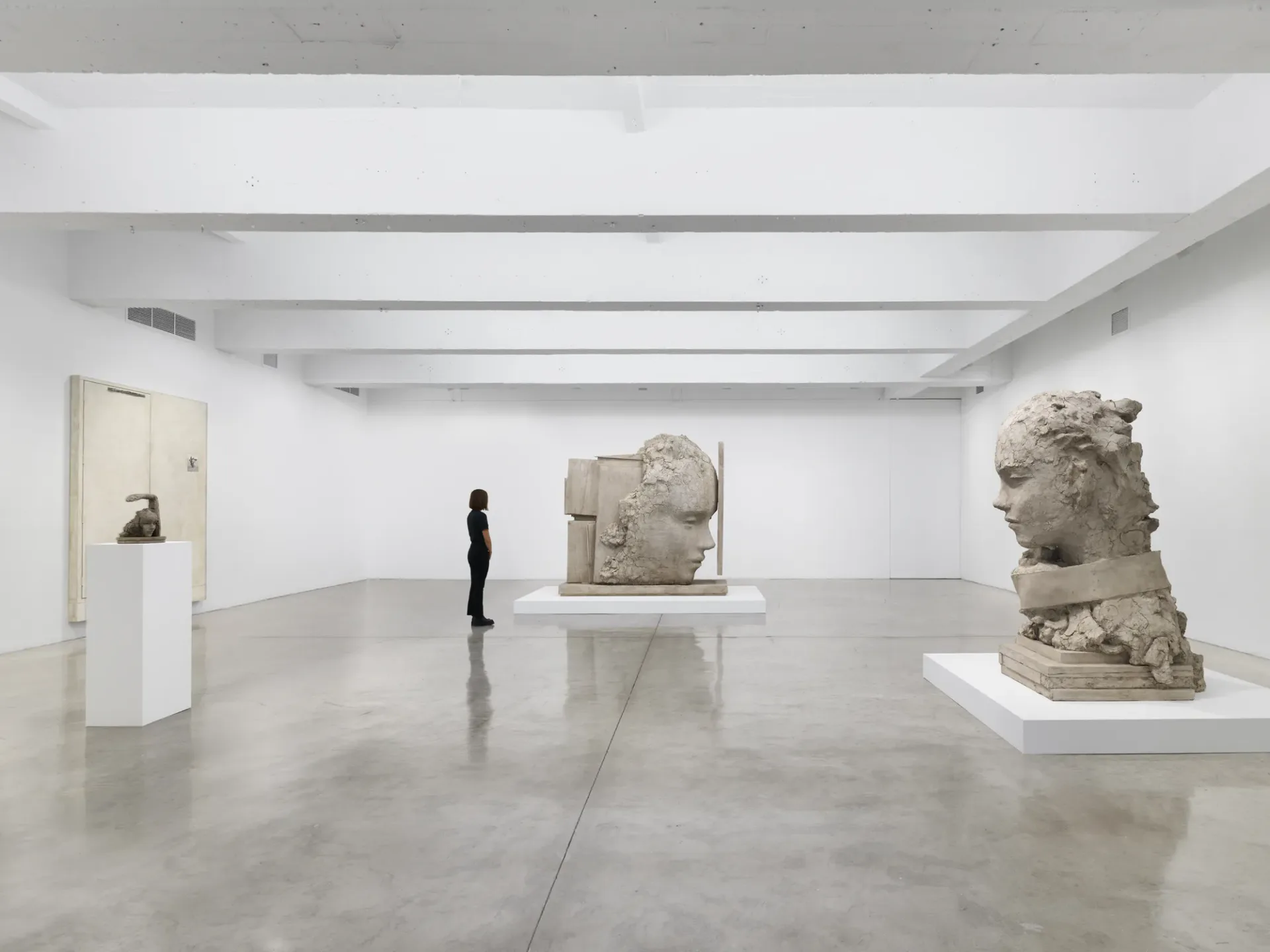 Gallery space with three limestone or sandstone sculptural heads displayed on white pedestals against clean white walls.