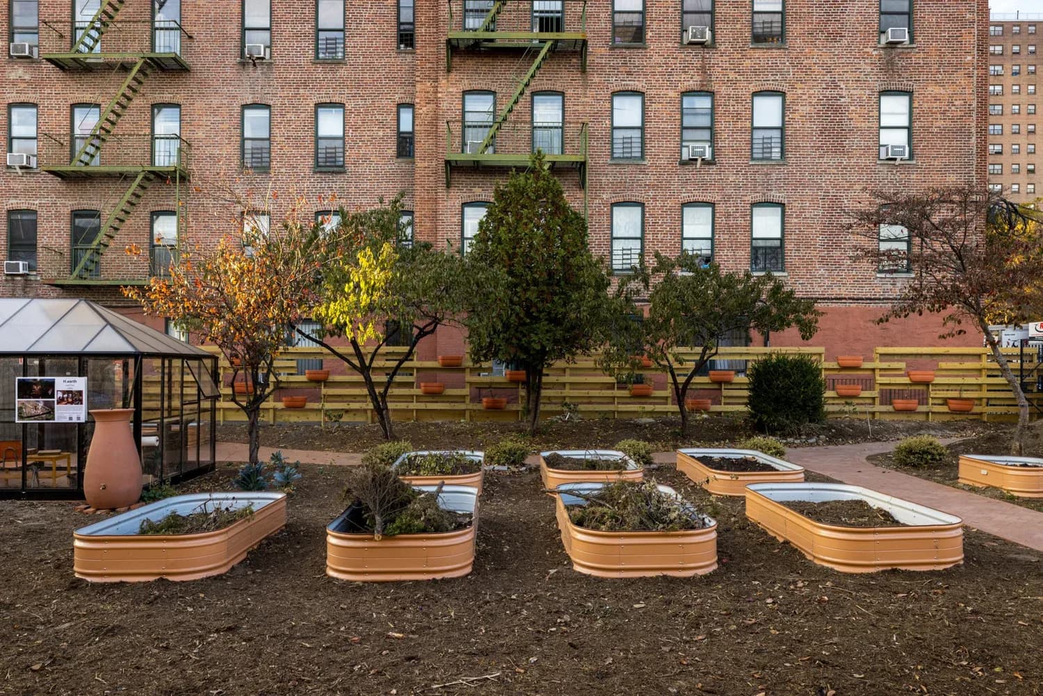 Urban community garden with wooden raised planting beds in front of a brick apartment building, autumn foliage visible.
