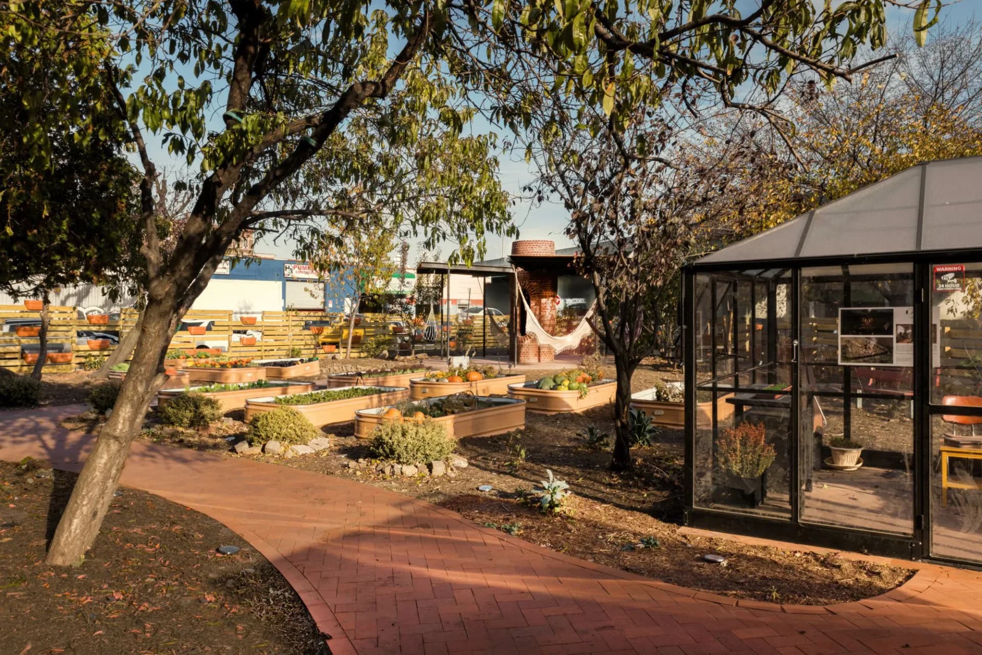 Outdoor garden space with mature trees, wooden structures, and a modern black-framed greenhouse or building, reddish paved pathway.