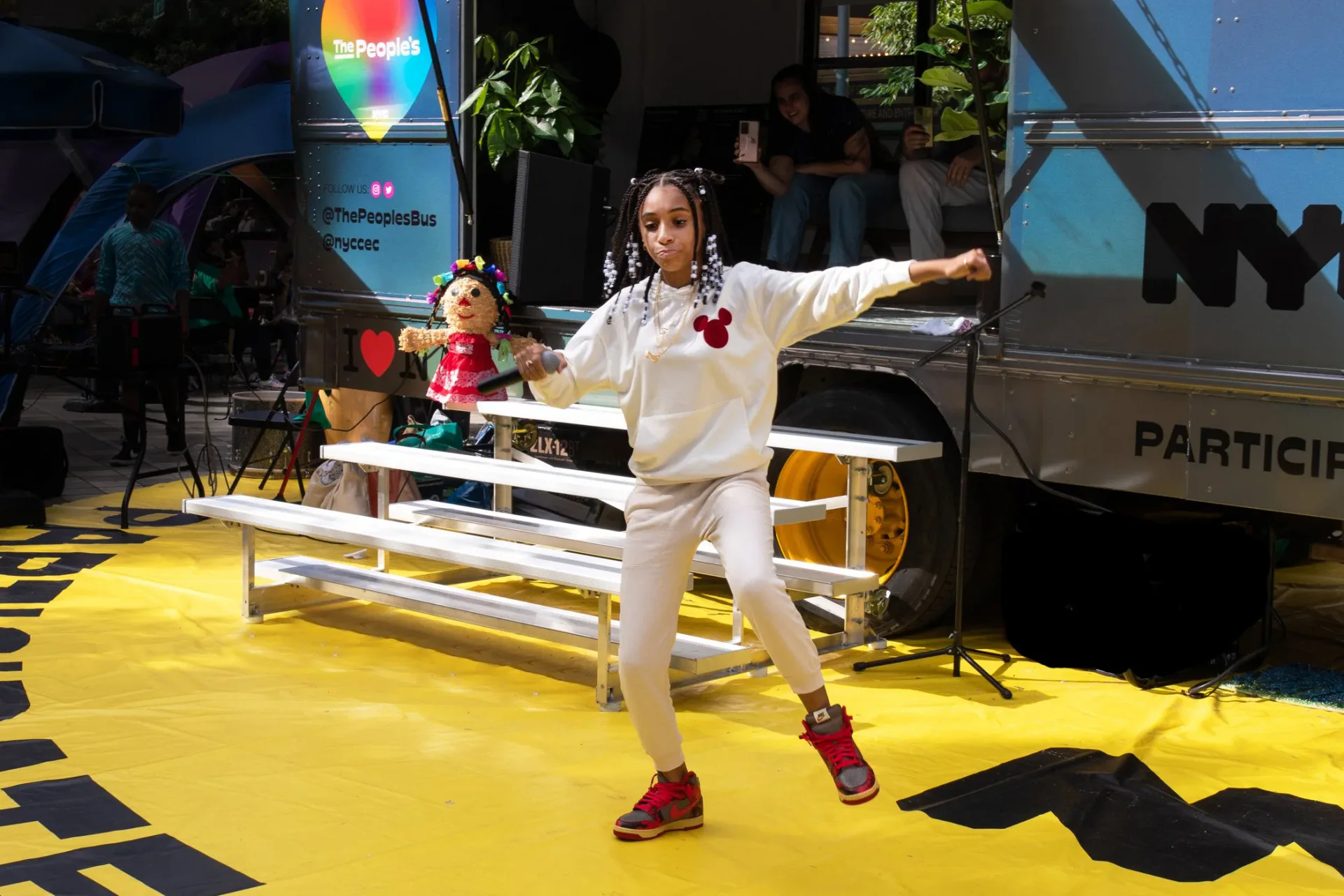 Performance installation with person in white suit dancing on bright yellow platform with rainbow light projection and bus structure.
