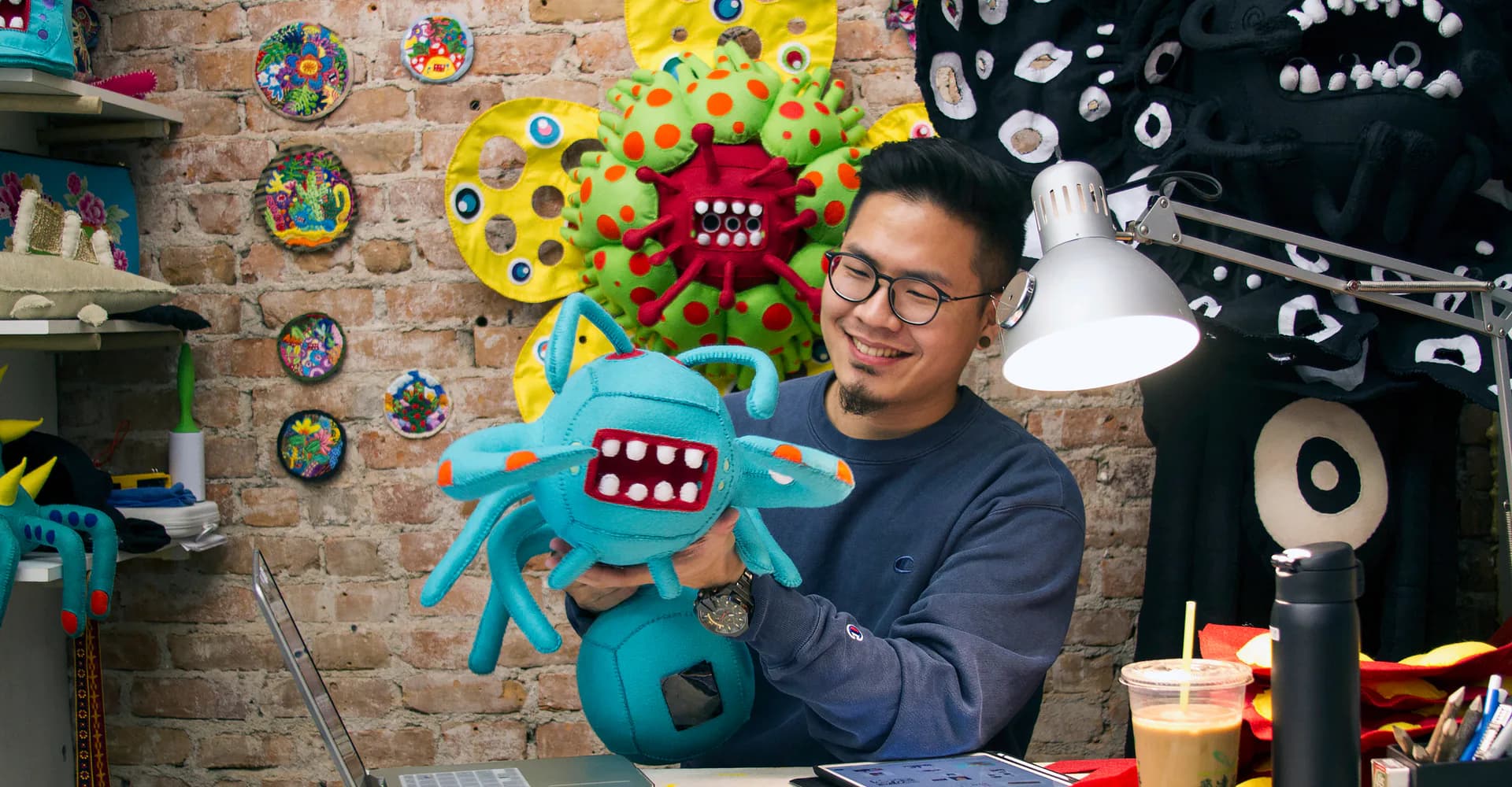 Person with glasses smiling while holding a turquoise sculptural creature with red features in a studio with brick wall and fiber works.