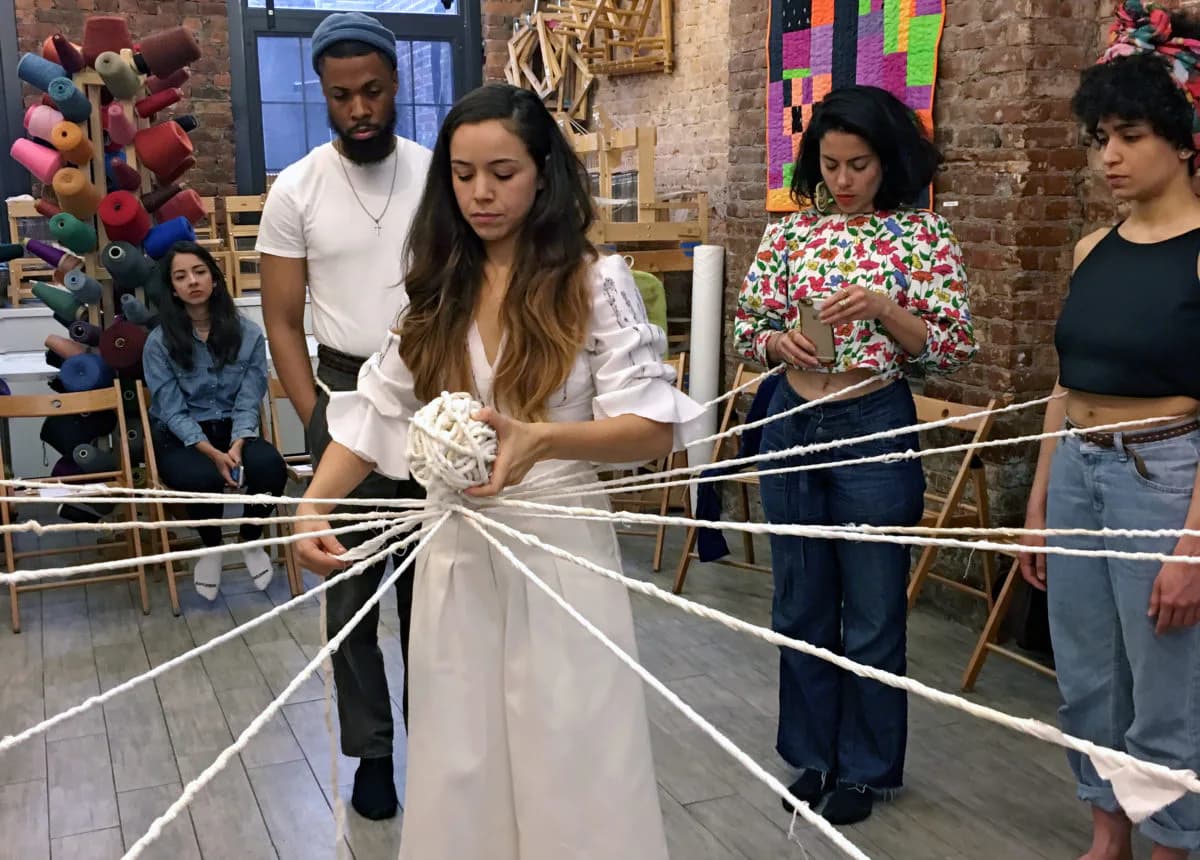 Group of people gathered around a large-scale fiber artwork installation with extended threads in a brick gallery space.