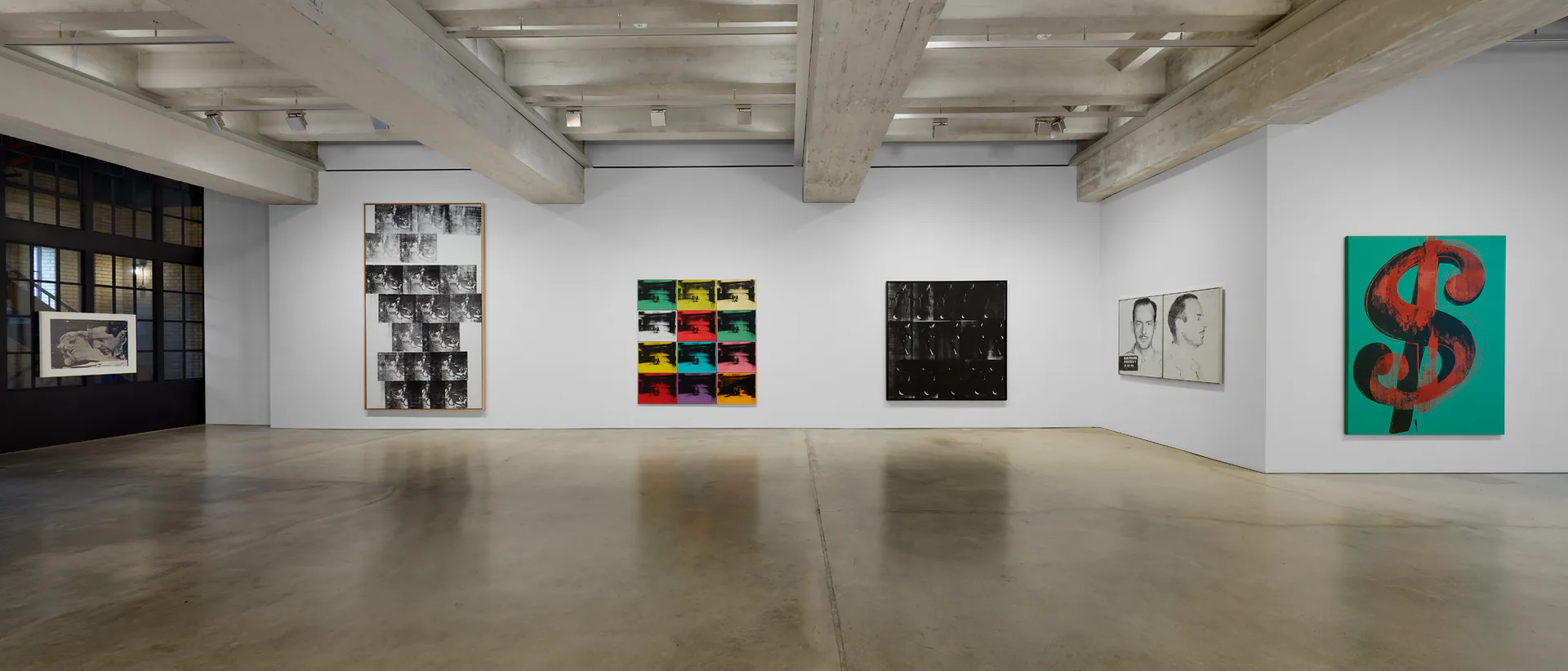 Contemporary gallery space with polished concrete floor, white walls, and exposed ceiling beams displaying an array of framed artworks including portraits, abstract compositions, and pop art pieces.