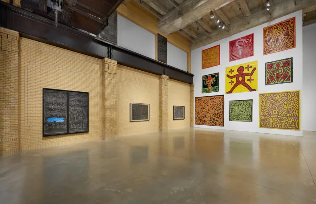 Gallery interior with exposed wood beam ceiling, brick wall on left, and white walls on right displaying colorful grid of abstract artworks and a yellow Chinese character piece.