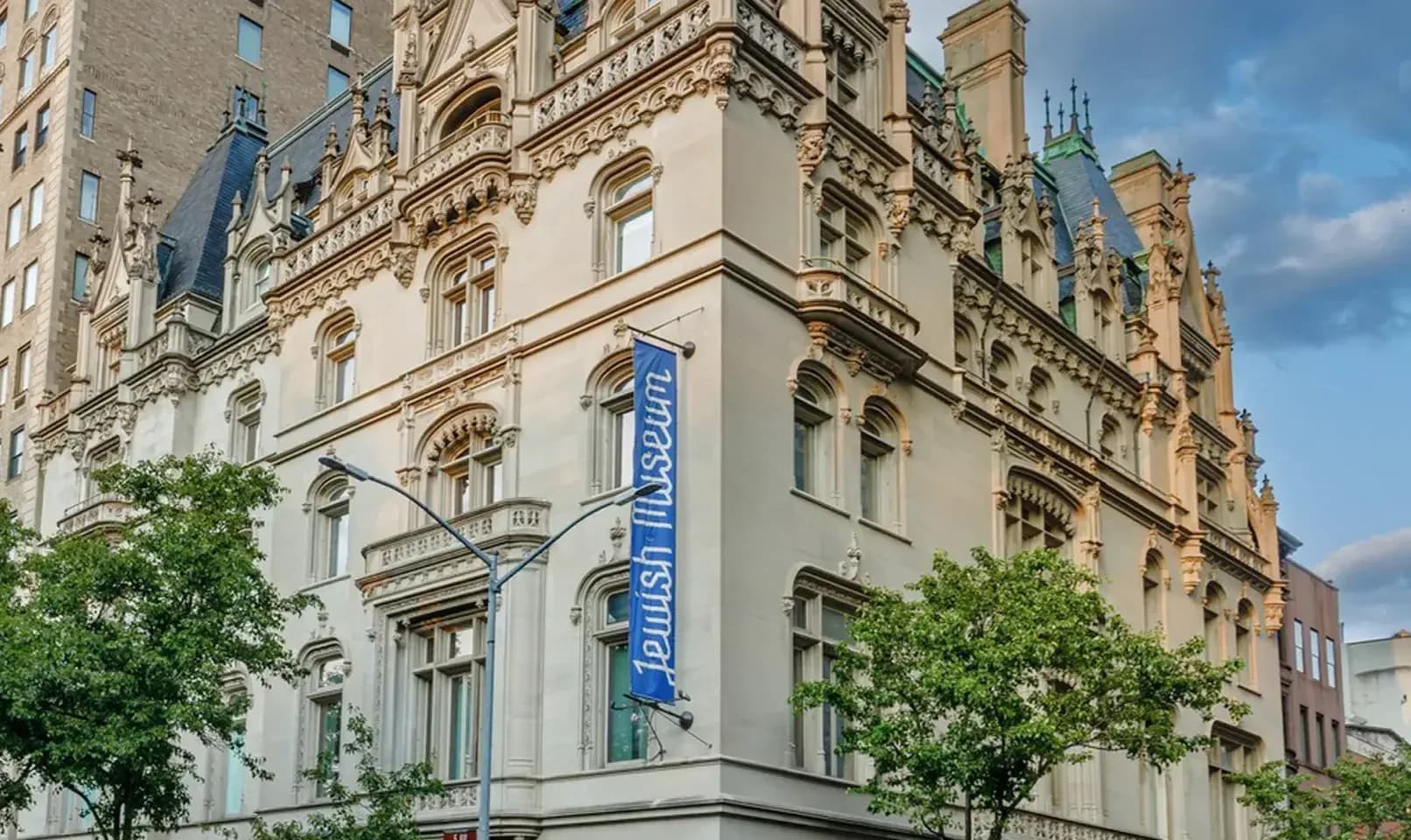 Exterior of a historic beige stone building with ornate architectural details, a blue banner hanging on the corner, and trees in foreground.