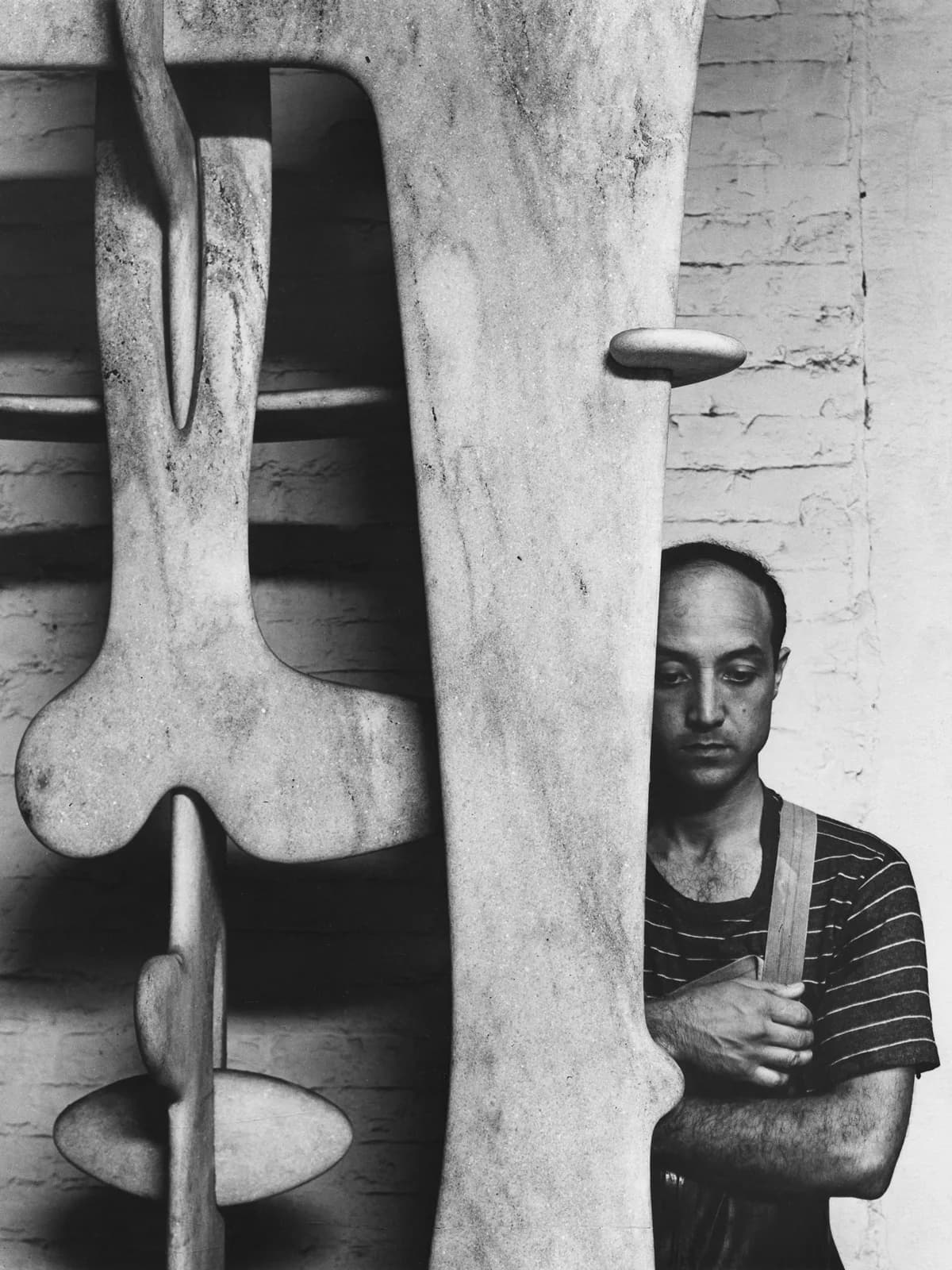 Black and white photograph of a man in striped shirt standing beside a large abstract sculptural form with organic curved shapes.
