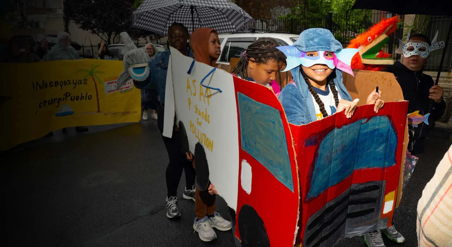 Group of children with umbrellas standing behind a handmade red and blue cardboard vehicle or art installation outdoors.