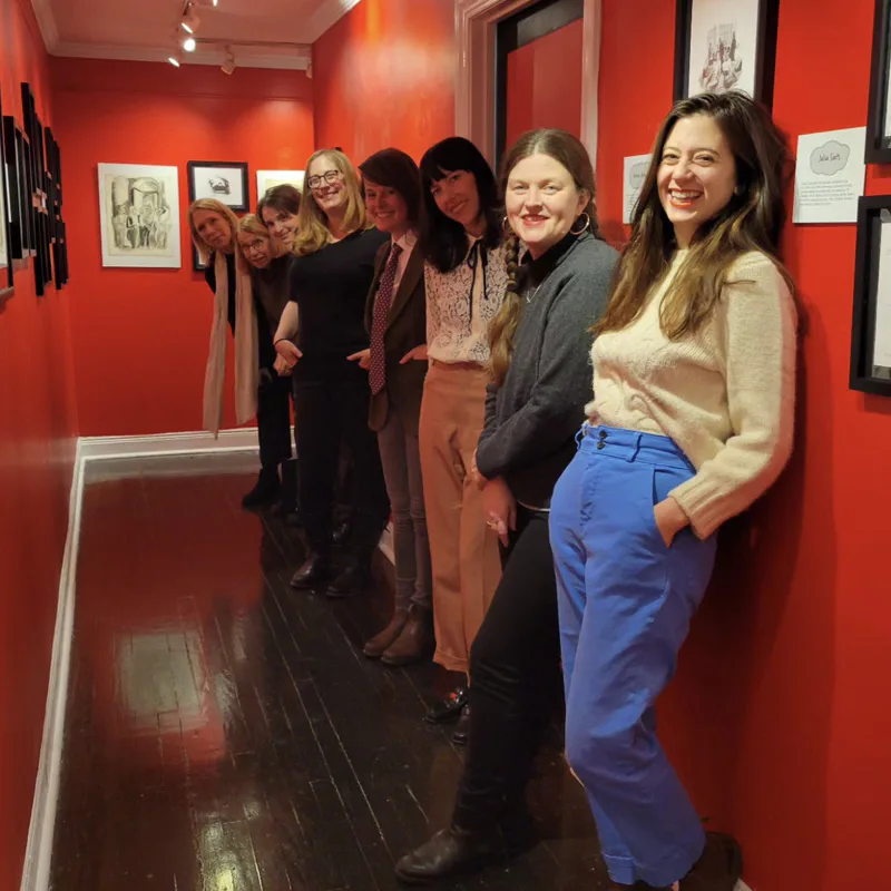 Group of six people standing in a gallery with bright red walls, framed artwork, and black flooring, smiling at the camera.