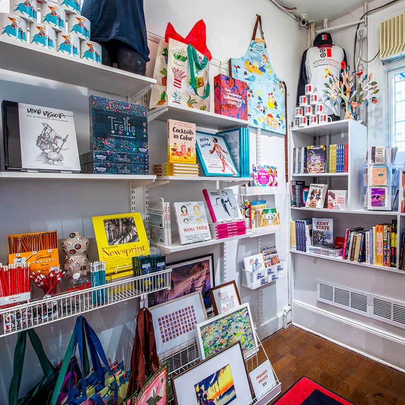 Wall-mounted white shelving displaying colorful children's books, illustrated prints, decorative items, and a large red bow, photographed indoors with natural light.