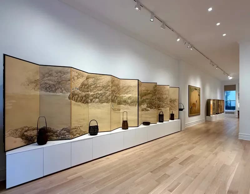 Gallery interior with a long folding screen painting displayed on white cabinets, wooden floors, and track lighting along a hallway