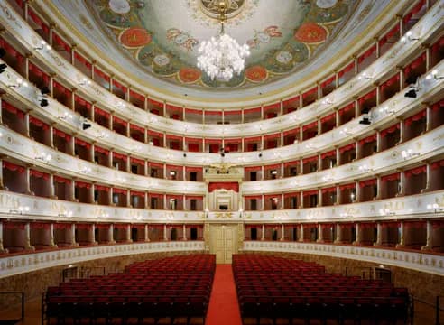 Interior photograph of ornate opera house with multiple balcony tiers, red velvet seats, gilt decoration, and crystal chandelier.