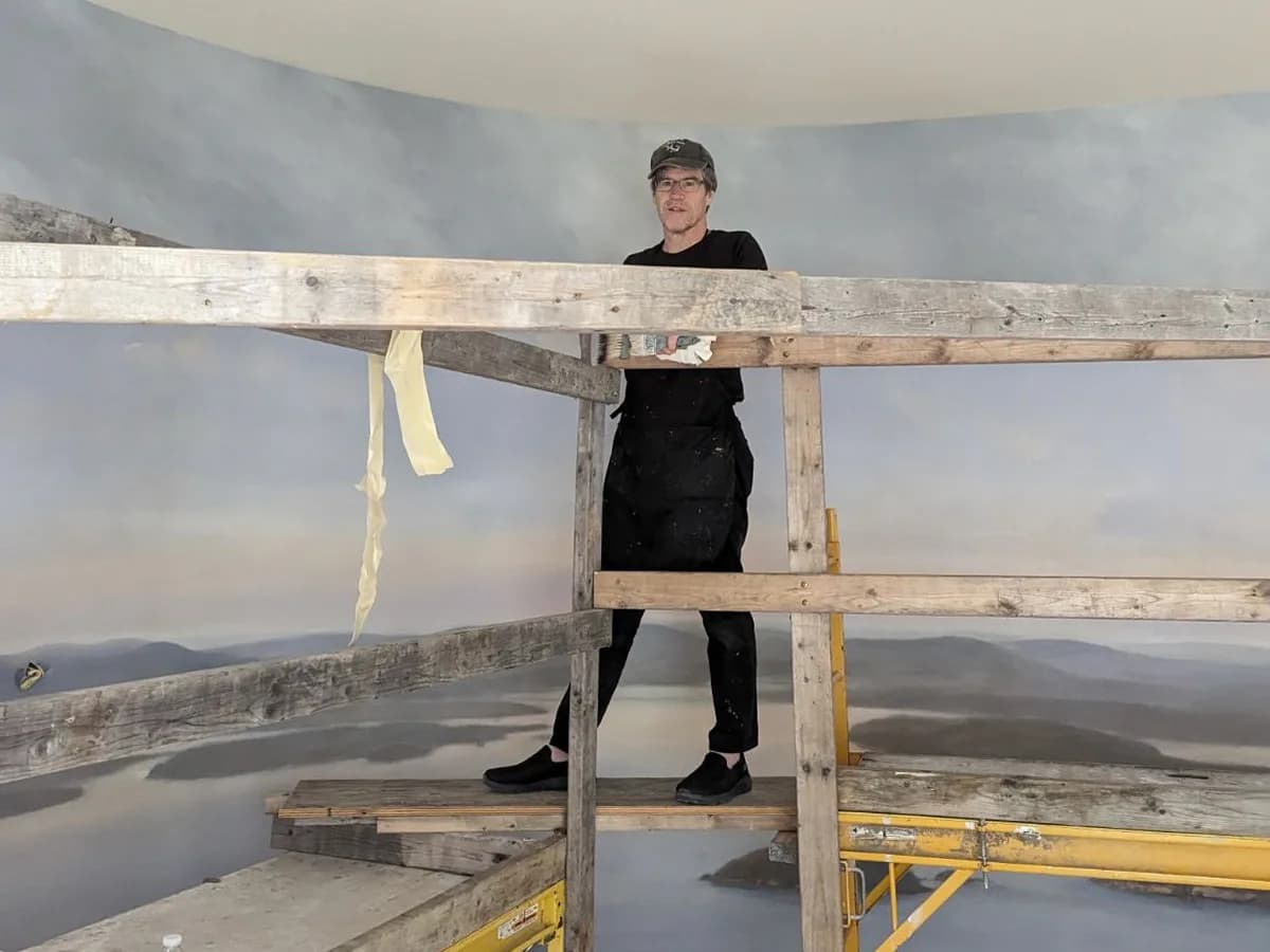 Person in black clothing positioned on wooden staging before a panoramic landscape mural installation in the studio space.
