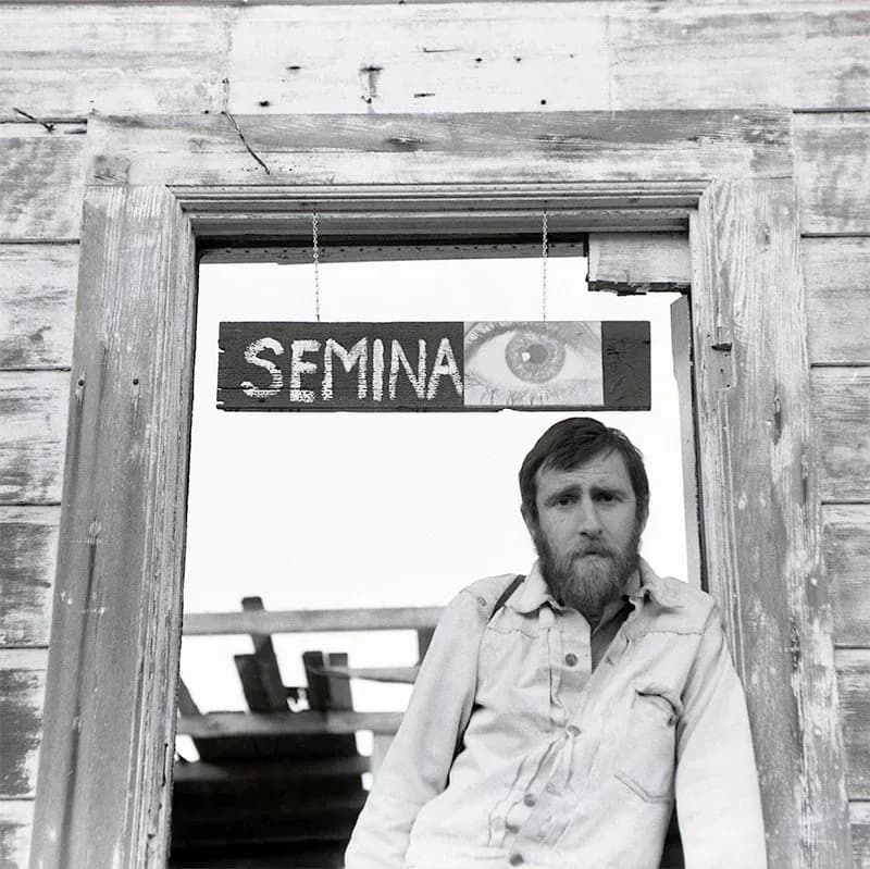 Black and white photograph of bearded man in light button-up shirt framed in wooden doorway with SEMINA sign visible above.