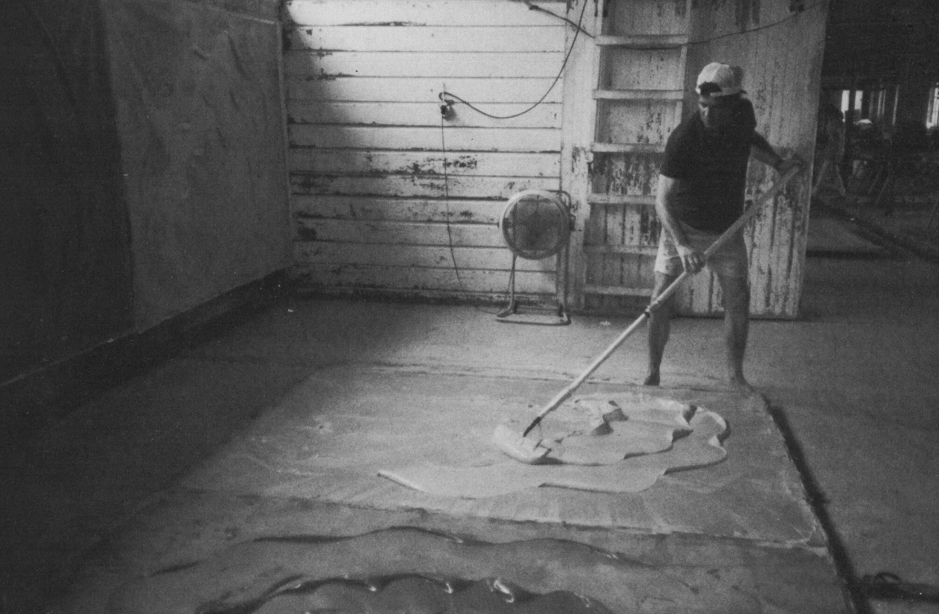 Black and white photograph of a person working on floor with tools in a sparse, industrial interior space with visible overhead wiring.