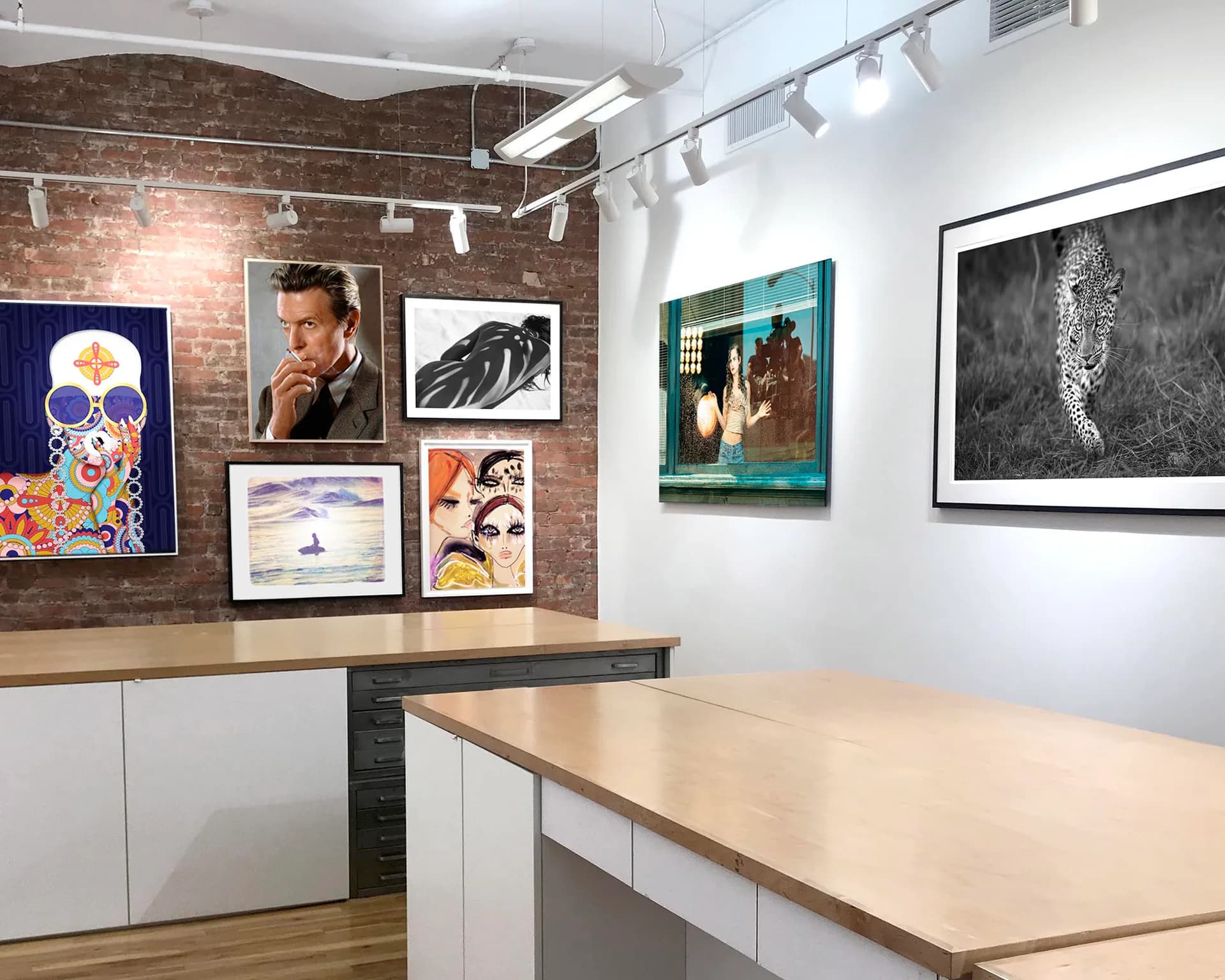 Gallery interior with exposed brick wall displaying colorful and black-and-white framed artworks, white counter and track lighting.