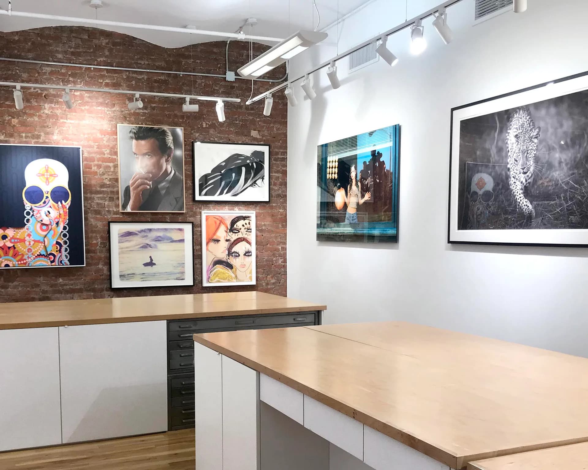 Gallery interior with brick wall displaying mixed-media framed artworks in various styles and colors above wooden counter with white cabinetry.