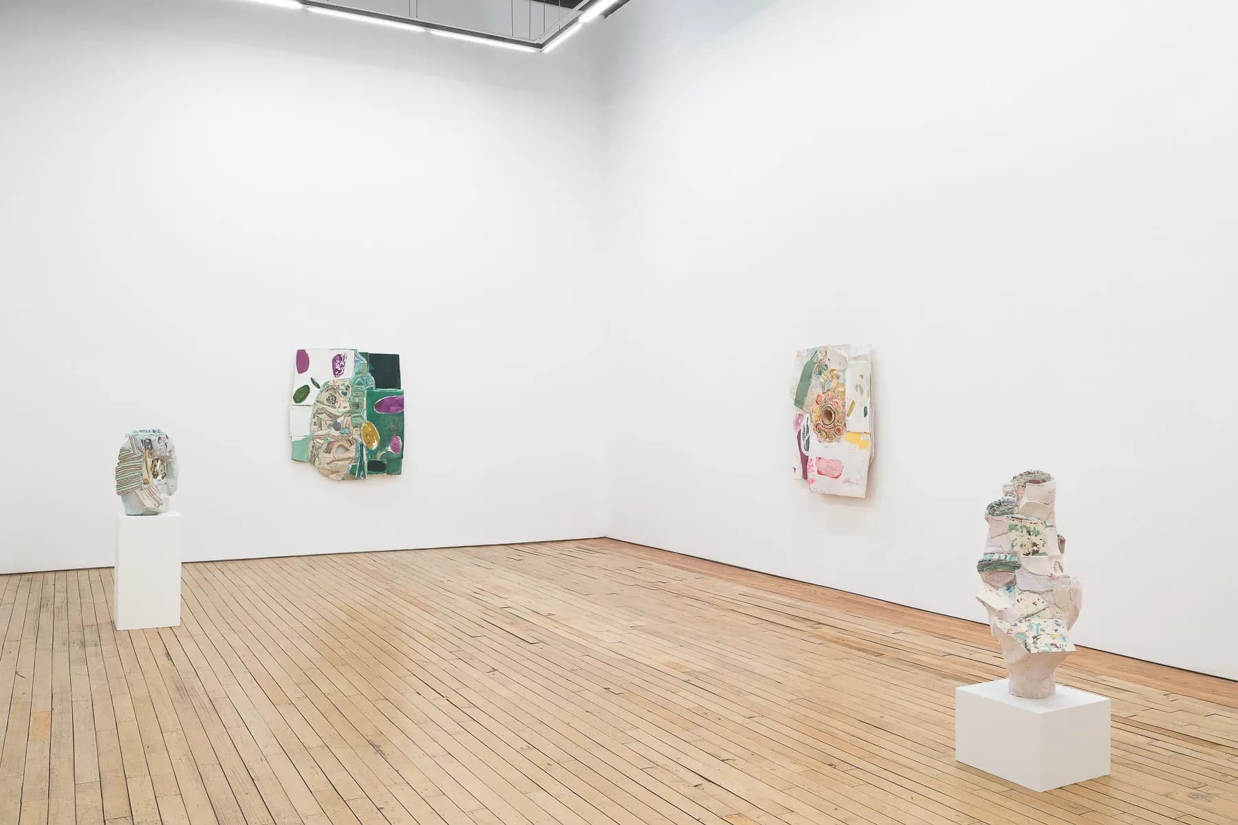 Gallery corner with white pedestals holding sculptural works and framed artworks on walls; wooden floor, recessed ceiling light.
