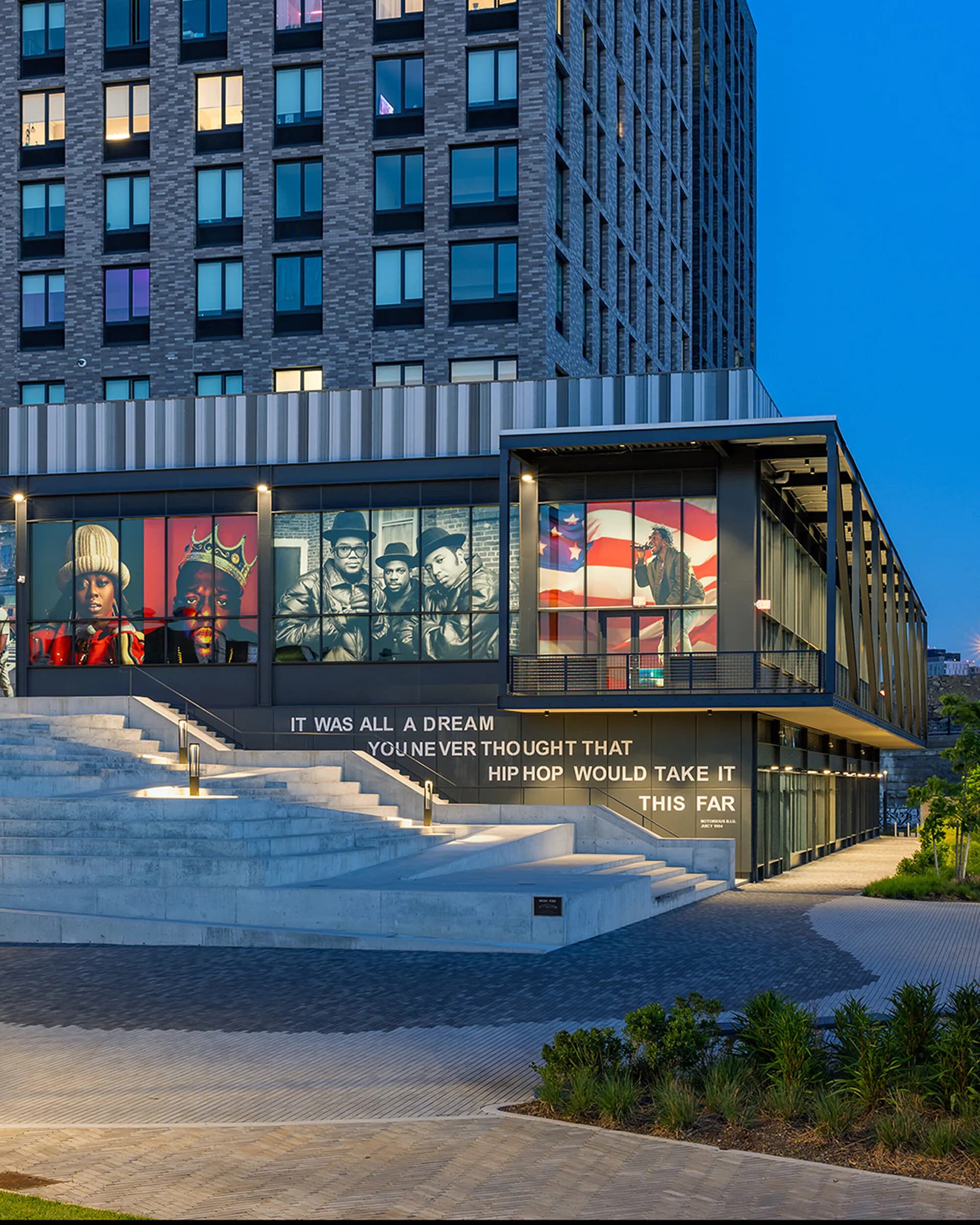 Exterior of Universal Hip Hop Museum building with illuminated windows, modern architecture, and ground-level exhibition windows displaying imagery and text about hip-hop history.