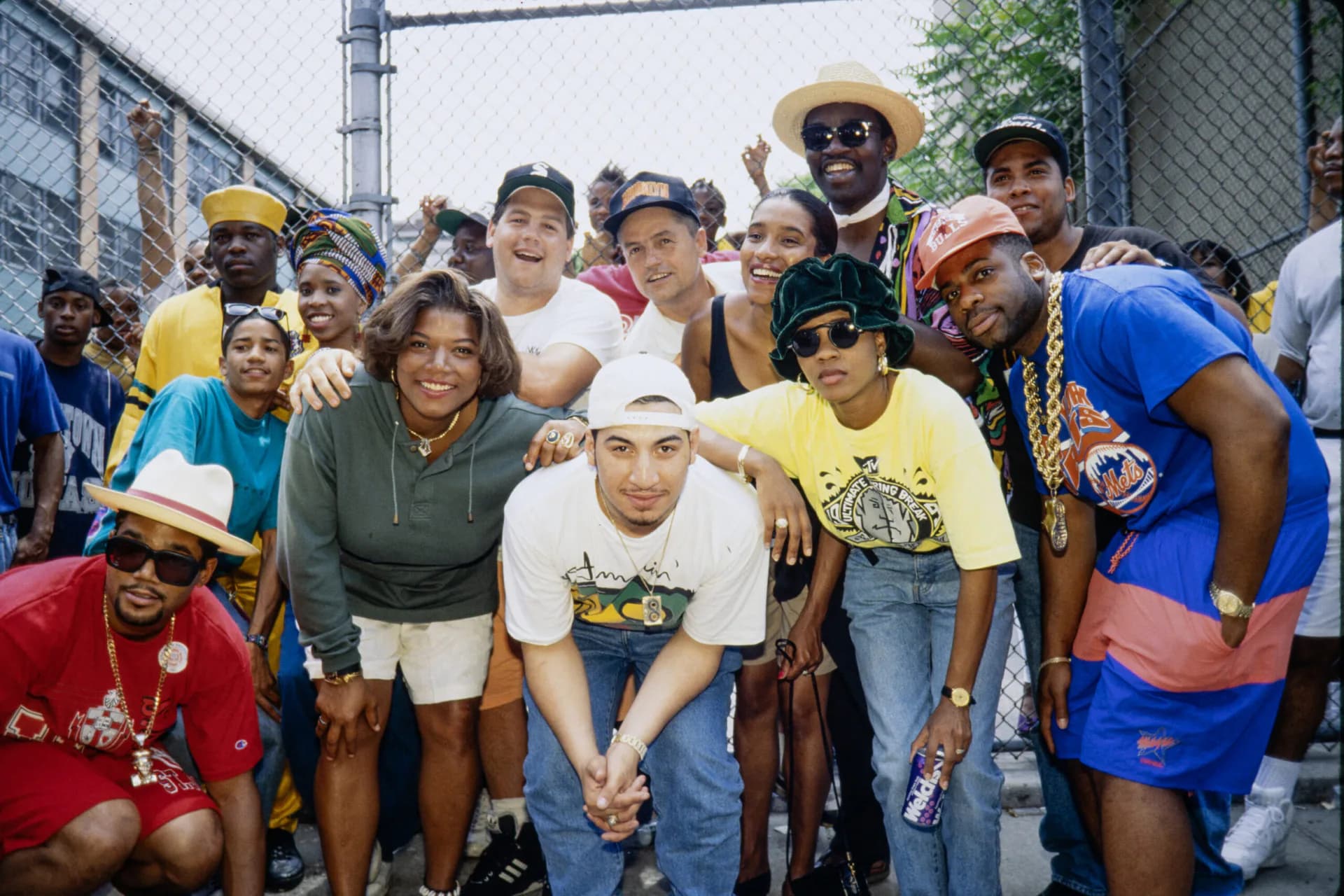 Color photograph of a diverse group of people gathered outdoors, many wearing baseball caps and casual clothing, smiling at the camera in a community street setting.