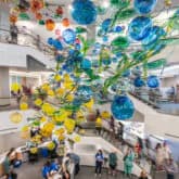 Interior of UrbanGlass with large suspended glass art installation featuring yellow and blue sculptural forms.