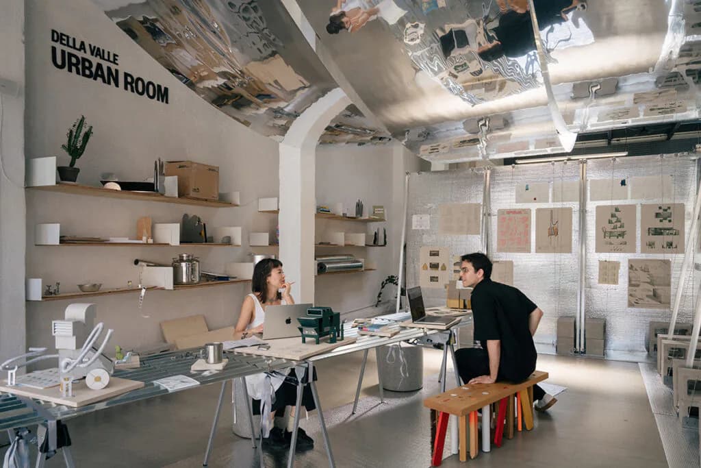 Photograph of Van Alen Institute's bright gallery interior with two people at desks, exposed ceiling, and shelving displaying architectural models and materials.