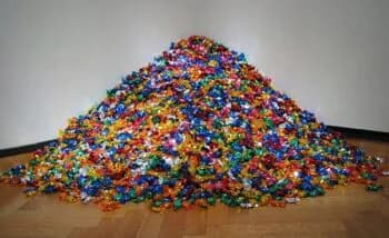 Colorful sculptural installation of thousands of small beads piled into a large mound on a wooden floor against white walls.