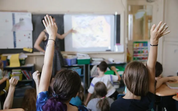 Classroom interior with students raising hands toward a projection screen, colorful bulletin boards and learning materials visible in background.