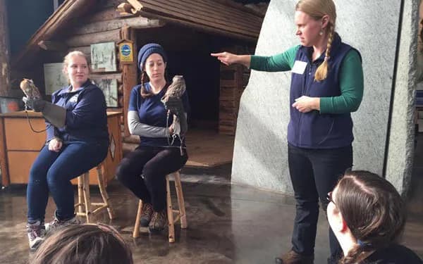 Interior workshop or classroom space with instructor gesturing during presentation to seated participants in blue vests.