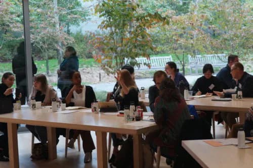 Long wooden table with seated panelists in covered outdoor pavilion overlooking green landscape, appearing to be a gallery or museum event space.