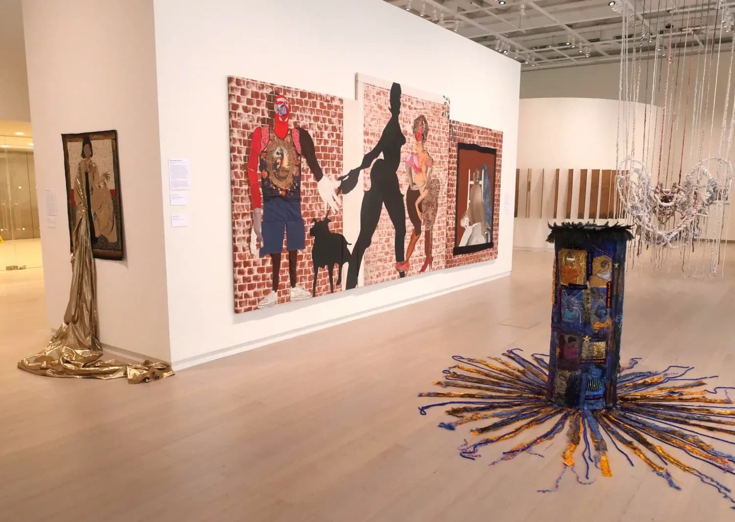 Interior of Wallach Art Gallery exhibition space with brick-patterned mixed-media wall installation, sculptural objects on floor, and industrial ceiling.