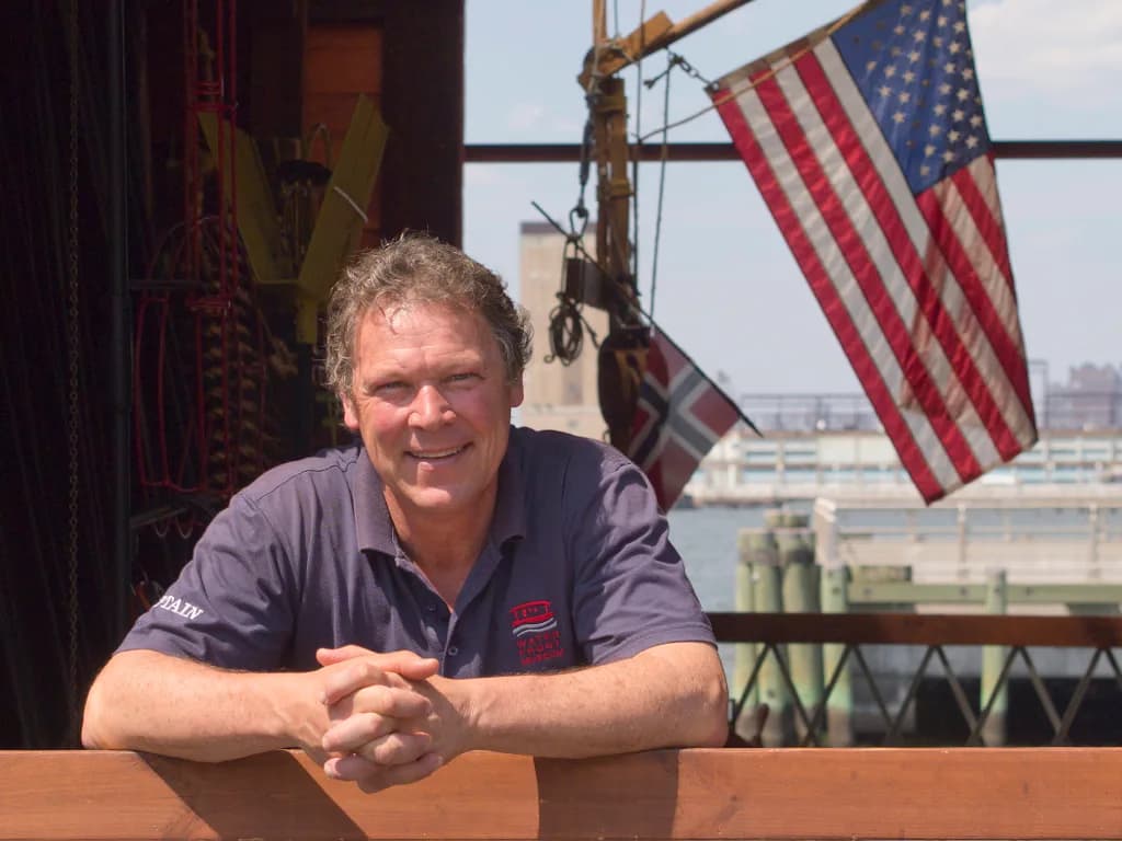 Man with gray hair in navy polo shirt seated at wooden table with American flags and waterfront industrial setting behind him.