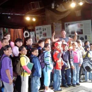 Group of schoolchildren gathered indoors at Waterfront Museum, wearing colorful jackets, viewing exhibits on white panels.