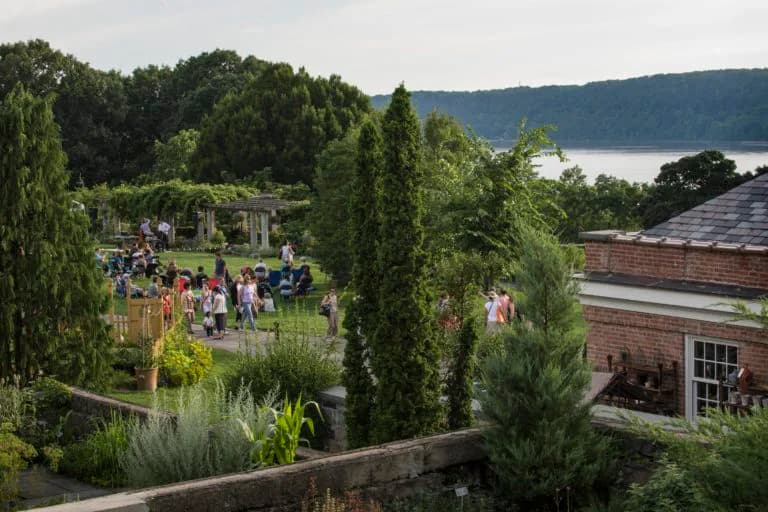 Elevated view of Wave Hill's manicured gardens with tall green columnar plants, red brick building, visitors walking paths, and distant water view.