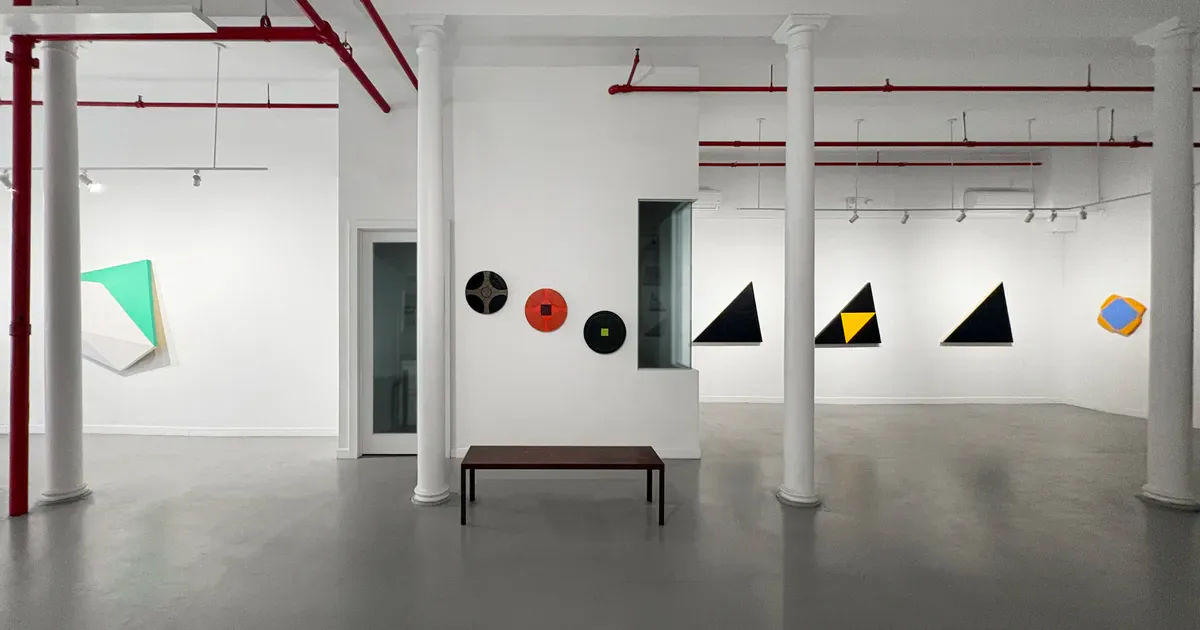 Westwood Gallery's minimalist white interior with red piping, displaying geometric paper artworks suspended from ceiling tracks and a brown bench.